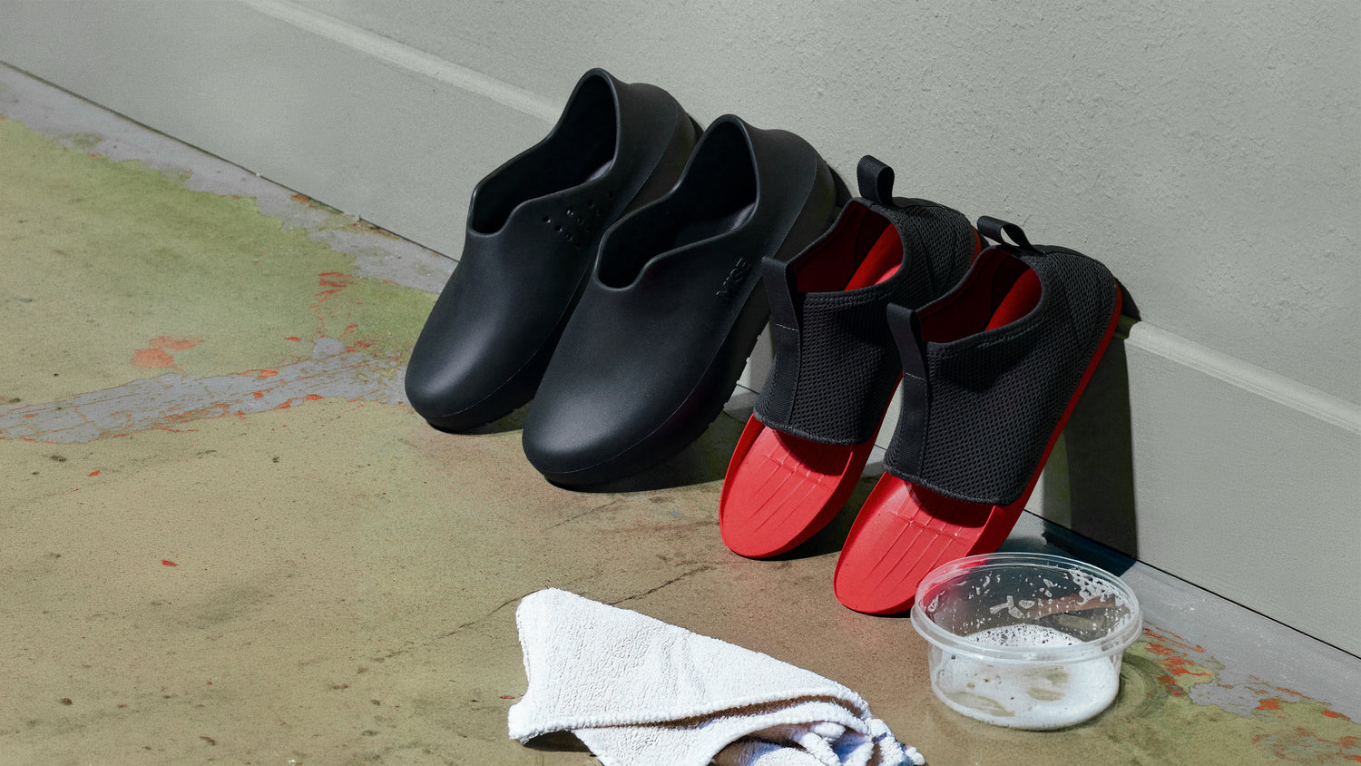 MISE All Day kitchen shoes with insoles removed arranged against a light wall on a concrete floor, with a white towel and a small clear plastic container filled with soapy water in the foreground